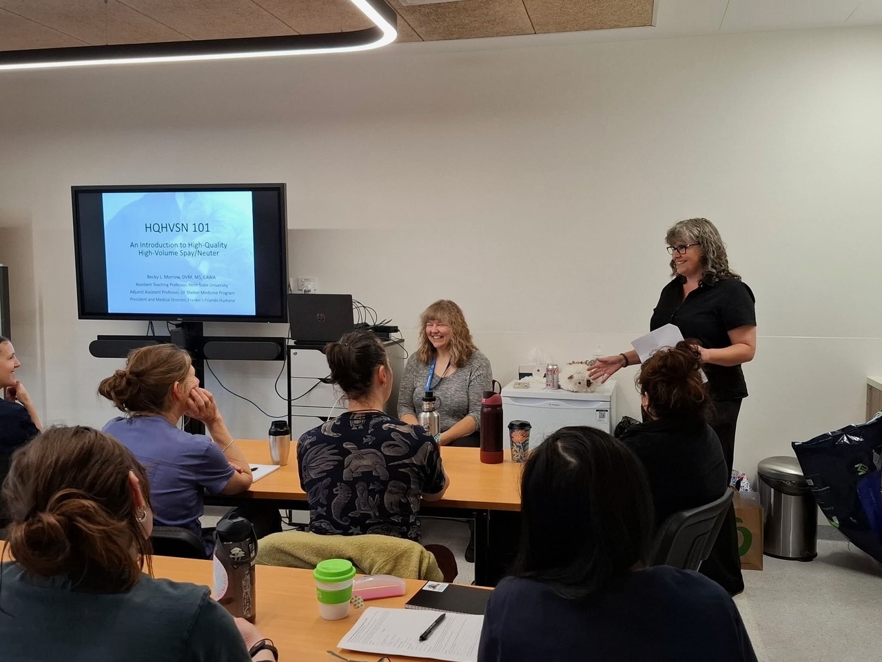 Workshop attendees seated around tables with two presenters beside a screen introducing high-quality, high-volume spay/neuter.